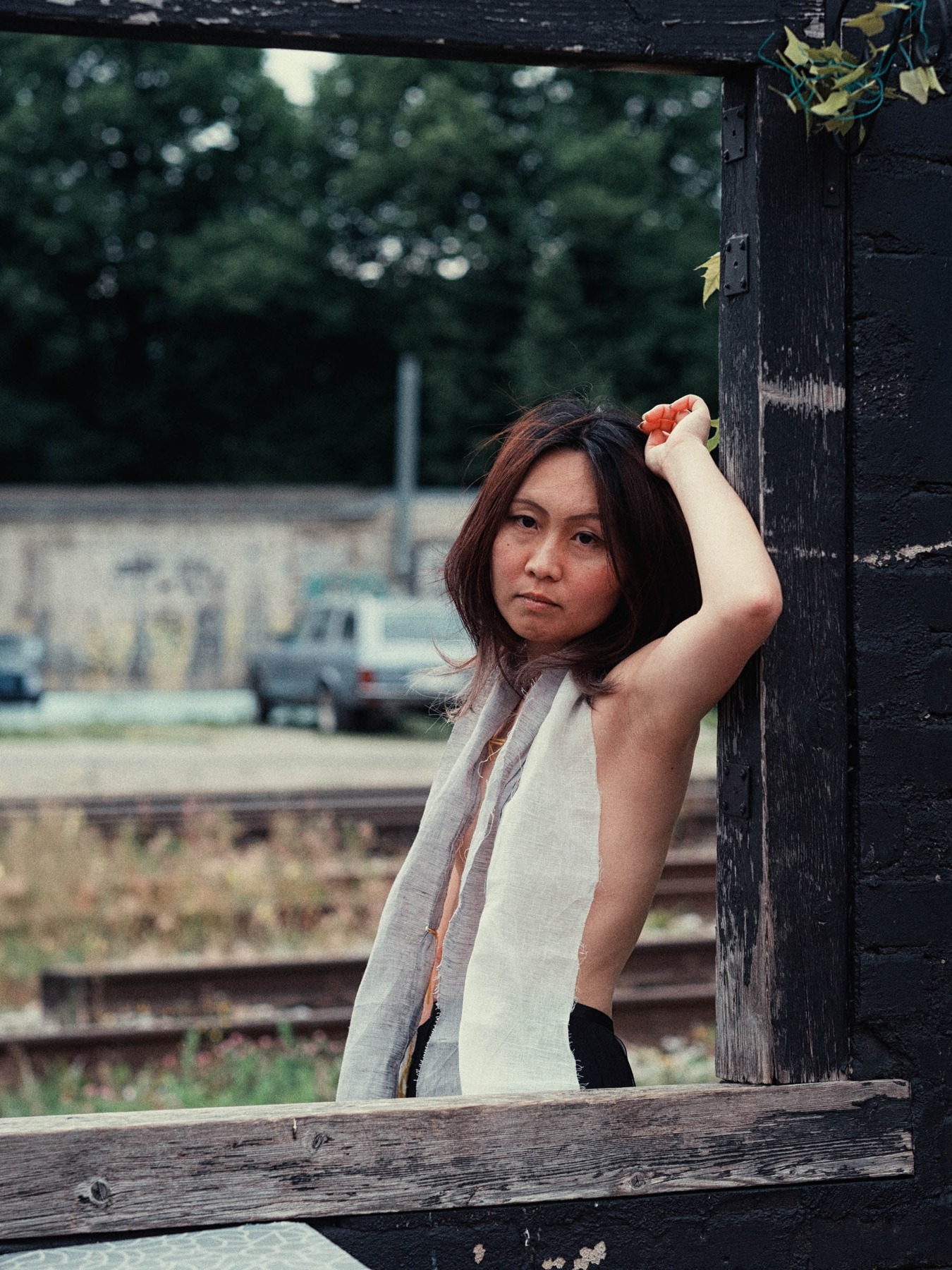 Lily-Yuri framed by weathered wood beside railway tracks in Tallinn, wearing a handmade linen garment