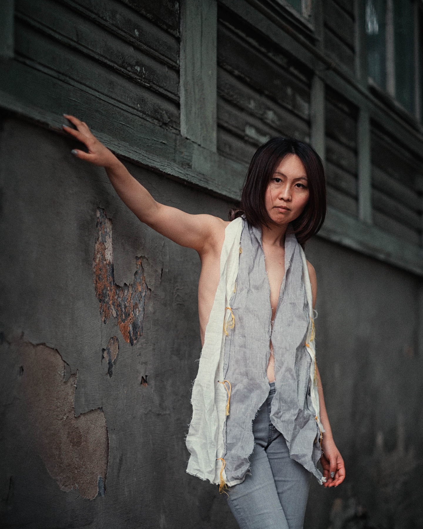 Lily-Yuri leaning against a dark wooden wall in Tallinn, wearing a draped linen garment